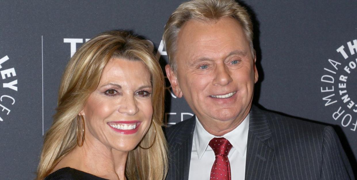 vana white and pat sajak pose for a photo and smile while looking past the camera, she is wearing large hoop earrings and a black top, he wears a gray suit jacket, white collared shirt and red tie with white polka dots