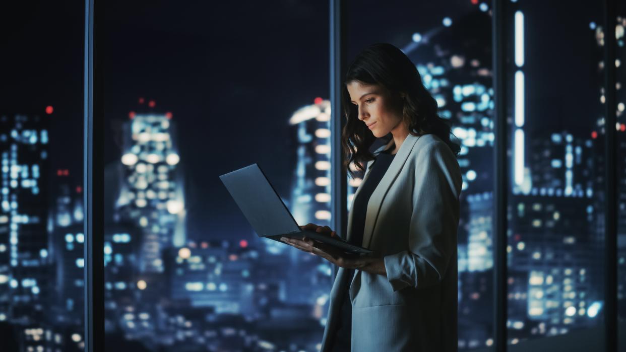  Woman standing in a room at night time with a backdrop of a city, while holding a laptop and using it with one hand . 