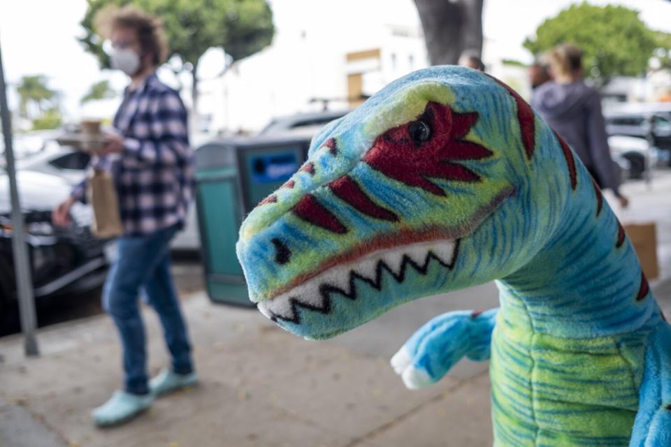 People are wearing masks and some are not wearing masks as they walk past a stuffed dinosaur