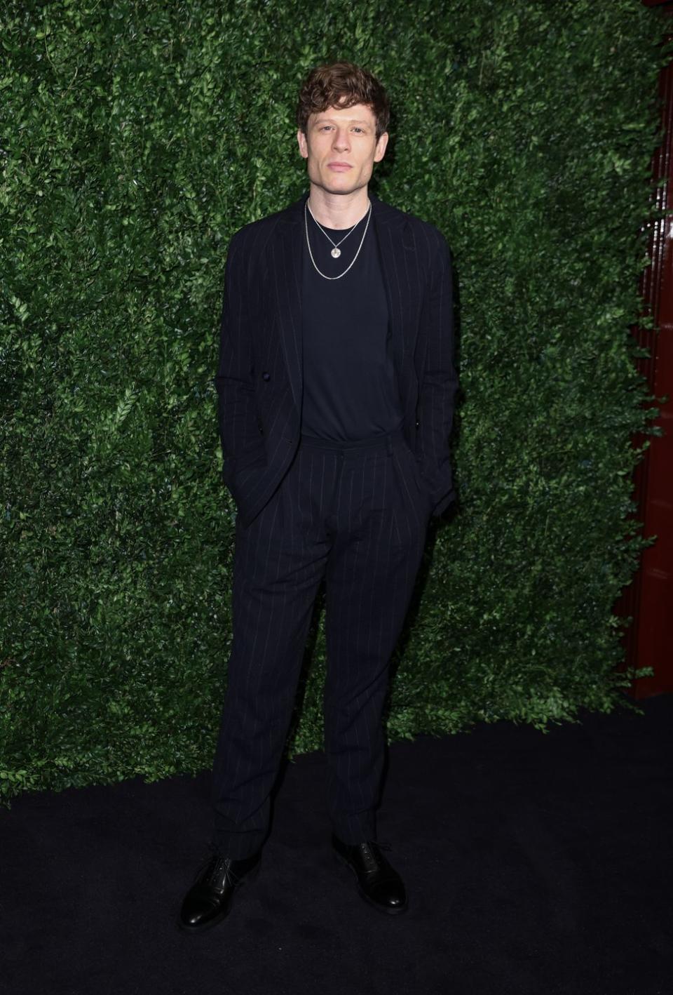 james norton wears a black suit and black top accessorised with two golden neck chains