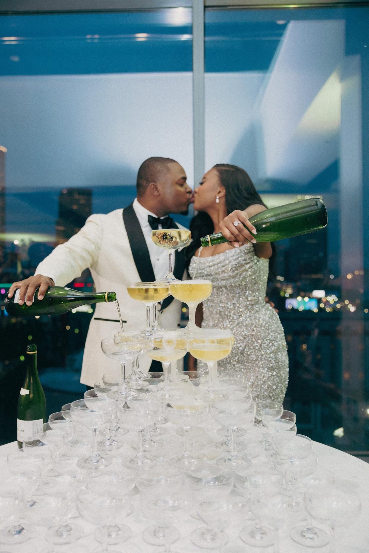 A bride and groom pour champagne into a tower of glasses while they kiss.