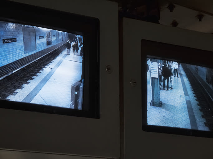 Two screens display security footage of people standing and walking on a subway platform