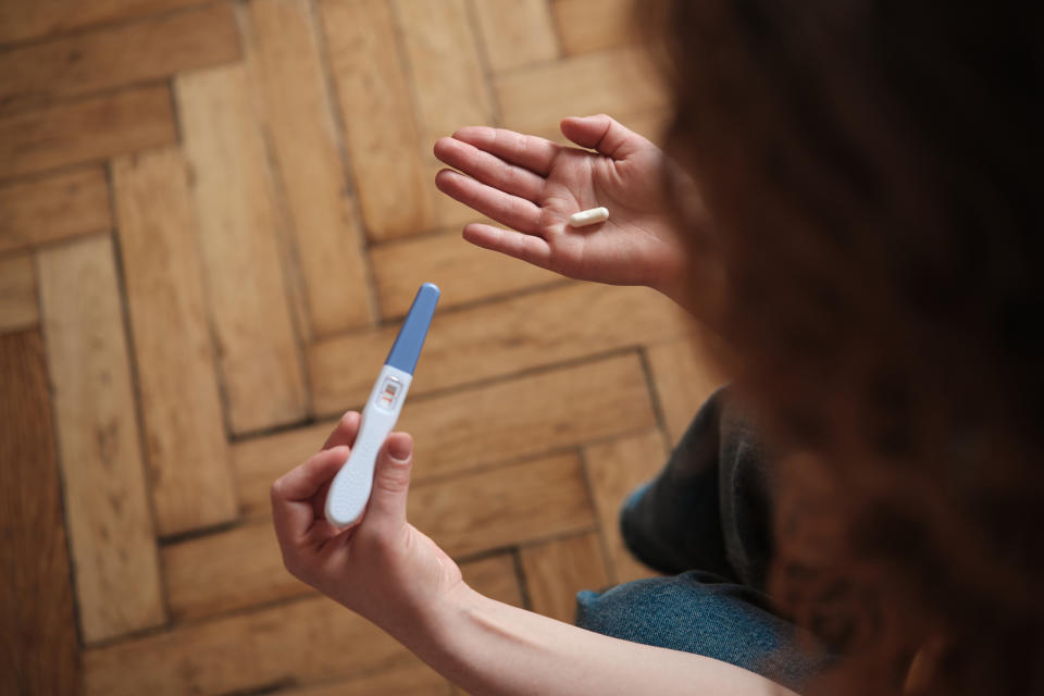 A person holds a pregnancy test in one hand and a single pill in the other, both above a wooden floor