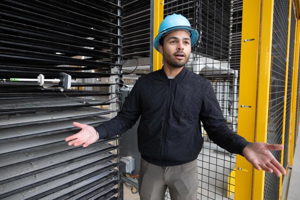 A man in a black jacket and blue hard hat stands beside a bank of trays