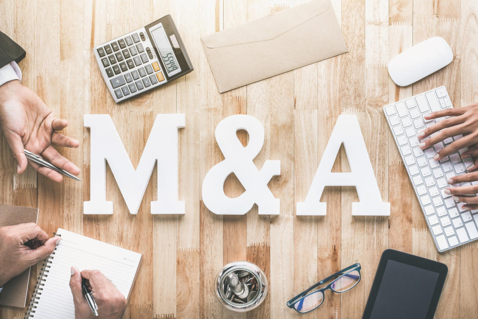 M&A spelled out with white block letters on a wooden table where people appear to be working.