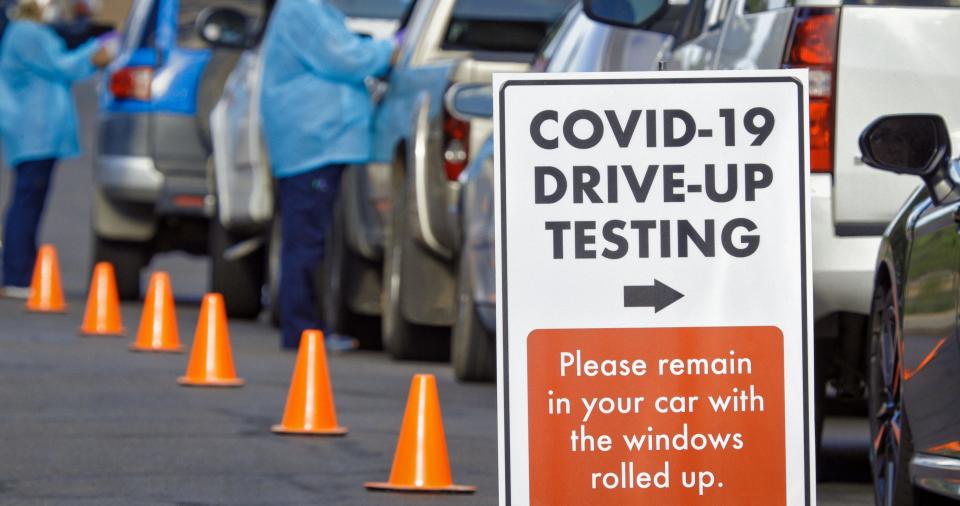 A "COVID-19 Drive-Up Testing" Sign Sits in the Foreground While Two Female Nurses Wearing Gowns and Surgical Face Masks Talk to Patients in their Cars in a Drive-Up (Drive Through) COVID-19 (Coronavirus) Testing Line Outside a Medical Clinic/Hospital Outdoors (Second Wave) in the Background