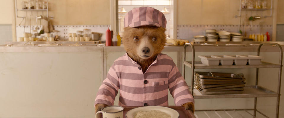 Paddington the bear dressed in pink and white prisoner uniform, holding a tray with a mug and bowl in a cafeteria setting from "Paddington 2"