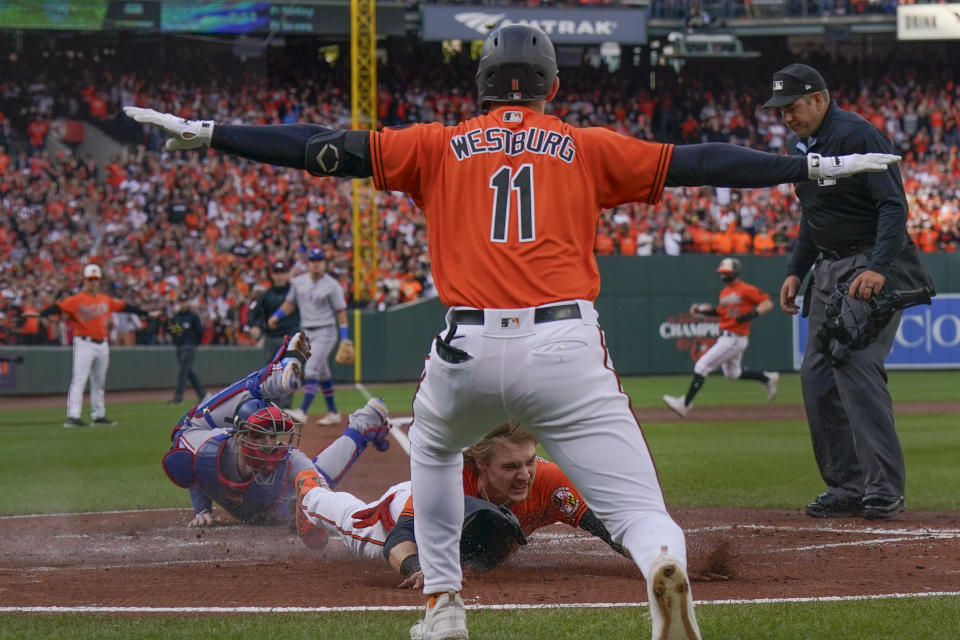 Baltimore Orioles' Jordan Westburg, top, signals safe as Gunnar Henderson, bottom center, scores past Texas Rangers catcher Jonah Heim, bottom left, on a single hit by Aaron Hicks during the first inning in Game 2 of an American League Division Series baseball game, Sunday, Oct. 8, 2023, in Baltimore. (AP Photo/Julio Cortez)