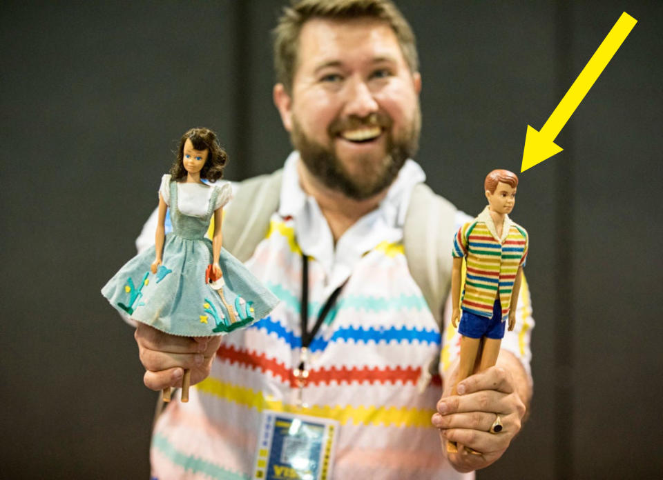 A man holds two vintage dolls—a female doll in a dress and a male doll in a striped shirt and shorts—smiling at the camera