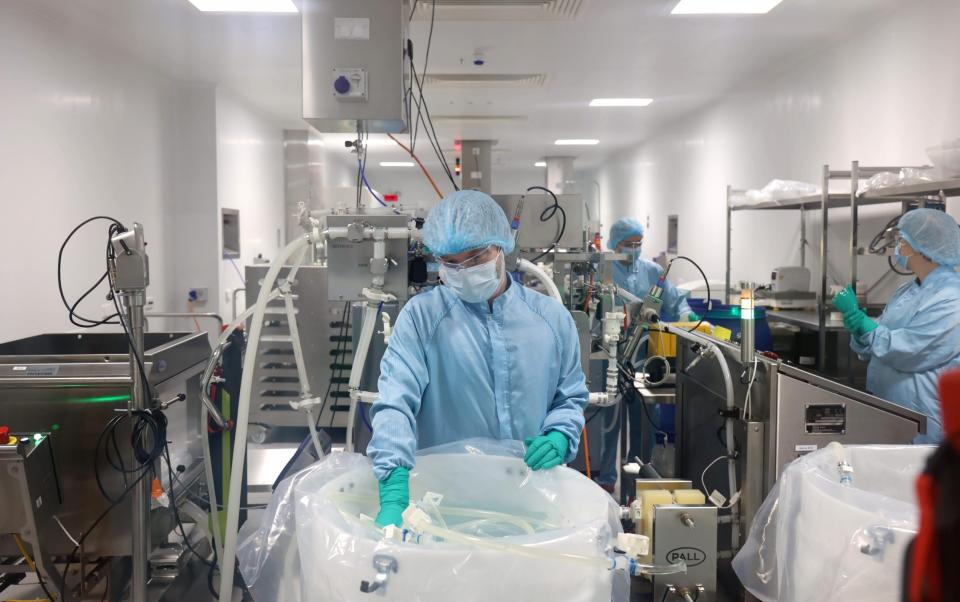 A scientist in a laboratory, in the ghost substance processing room, handles the equipment that produces the vaccine.  The worker, dressed in a blue gown and wearing a mask and rubber gloves, is surrounded by laboratory equipment