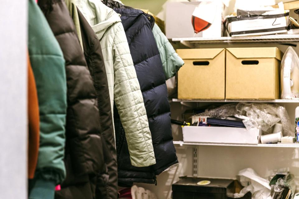 Cluttered coat closet with lots of coats, plus boxes and packages on a back shelf.