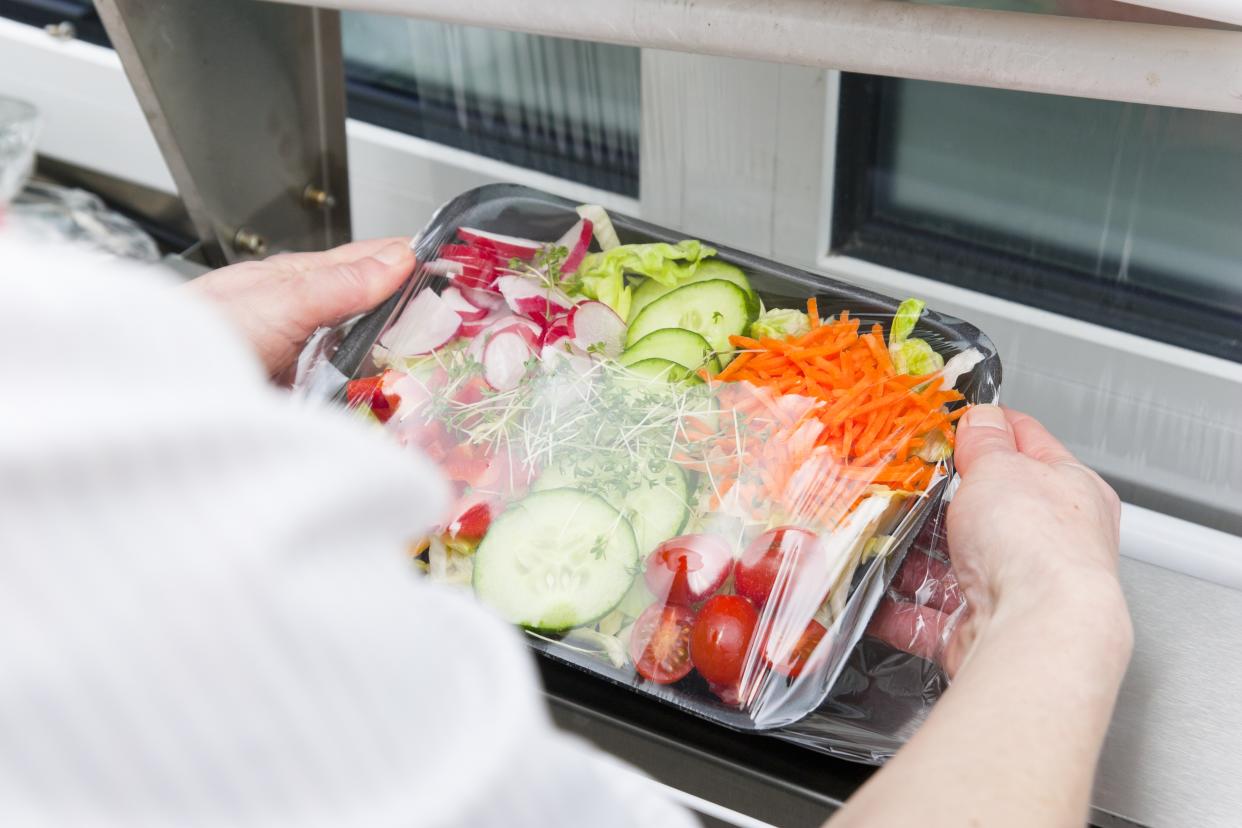 Production line of pre-packaged convenience food. Over the shoulder view, a woman wrapping ready to eat salad in a wrapping machine with cling foil. This image is part of a series.