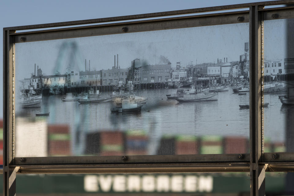 In this Tuesday, Oct. 19, 2021, photo containers stacked on an Evergreen cargo ship are seen through a historical picture of the Port of Los Angeles, part of the Terminal Island Japanese Fishing Village Memorial in San Pedro, Calif. California Gov, Gavin Newsom on Wednesday, Oct. 20, 2021 issued an order that aims to ease bottlenecks at the ports of Los Angeles and Long Beach that have spilled over into neighborhoods where cargo trucks are clogging residential streets. Last week the White House allowed the ports complex to become a 24-hour operation in an effort to break the logjam and reduce shipping delays. (AP Photo/Damian Dovarganes)