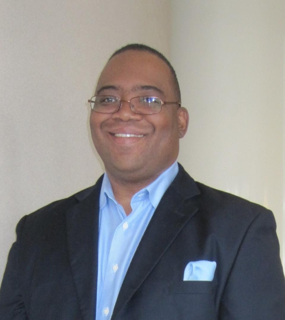 a man with close cut brown hair and glasses wearing a nice suit with a blue button up and matching blue pocket square, smiling at the camera