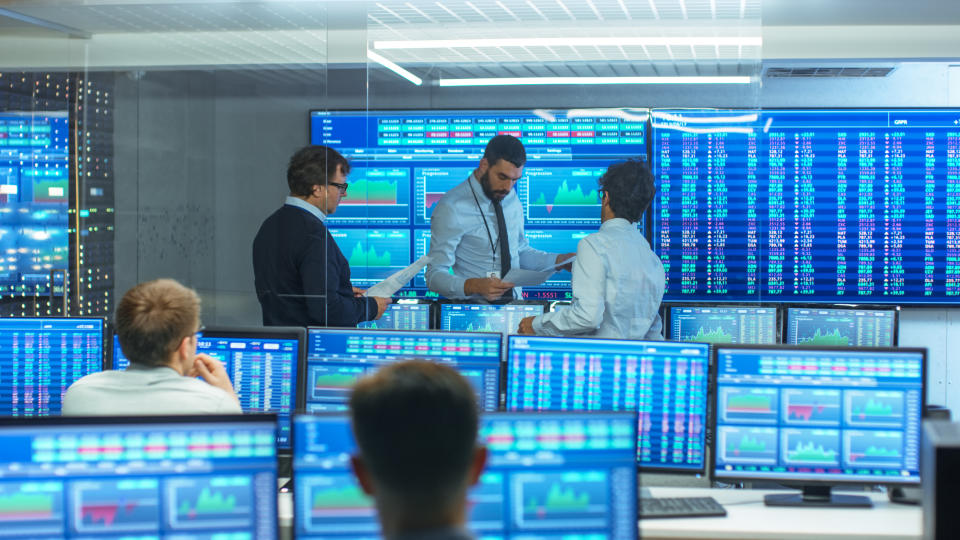 Three people talking at the front of a room with many monitors showing charts and numbers.