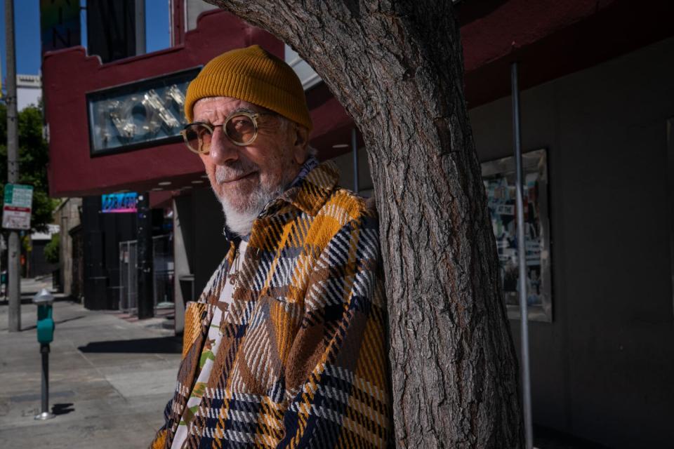 A fashionable older man in a plaid coat leans against a tree