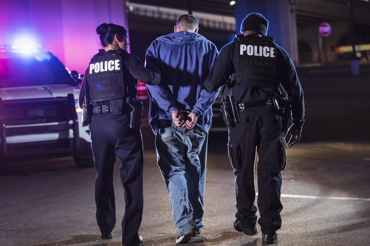 Rear view of two multiracial police officers arresting a criminal suspect, a man with his hands handcuffed behind his back. One of the officers is an African-American man and the other is a policewoman, both in their 40s.