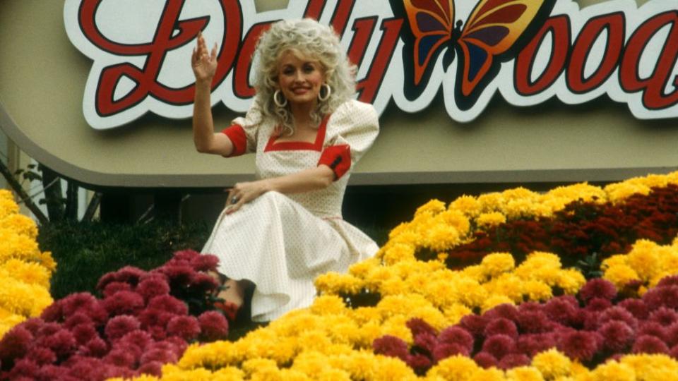 dolly parton wearing a white and red dress, sitting down in front of a large dollywood sign and waving, with several red and yellow flowers in front of her