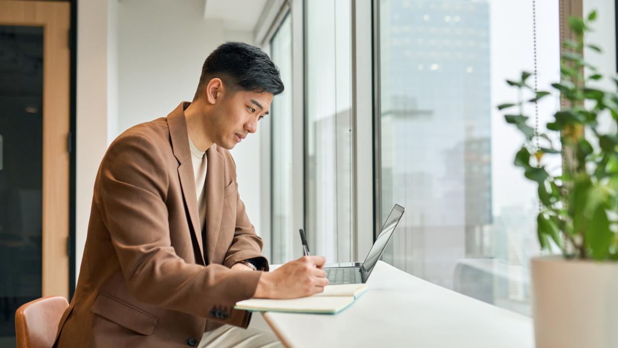  A young man working on laptop in office writing notes. 