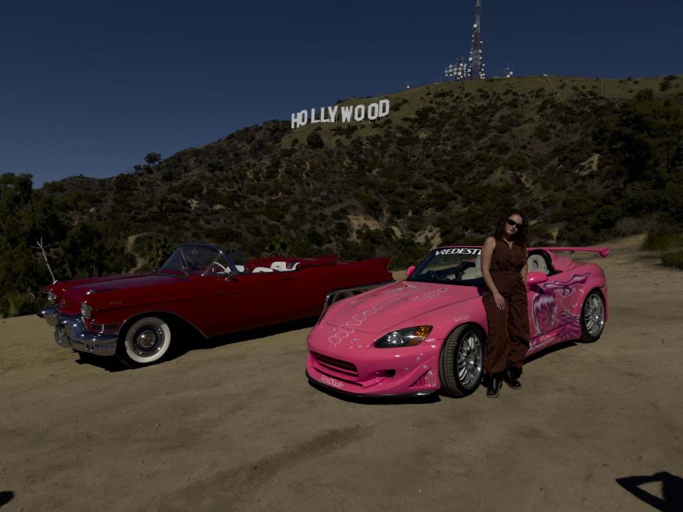 a person standing next to a pink car in front of a hill