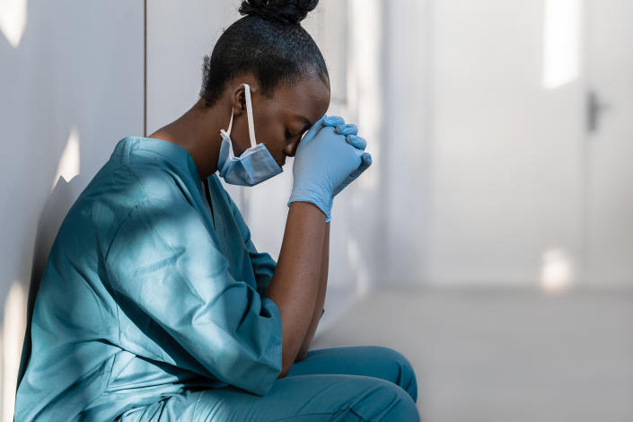 Tired depressed female doctor wears face mask blue uniform gloves sits on hospital floor. (Getty Images)