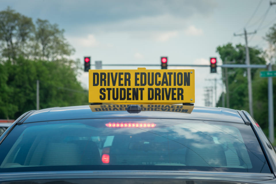 A Driver's Education car with a "Student Driver" sign on top is stopped at a traffic light