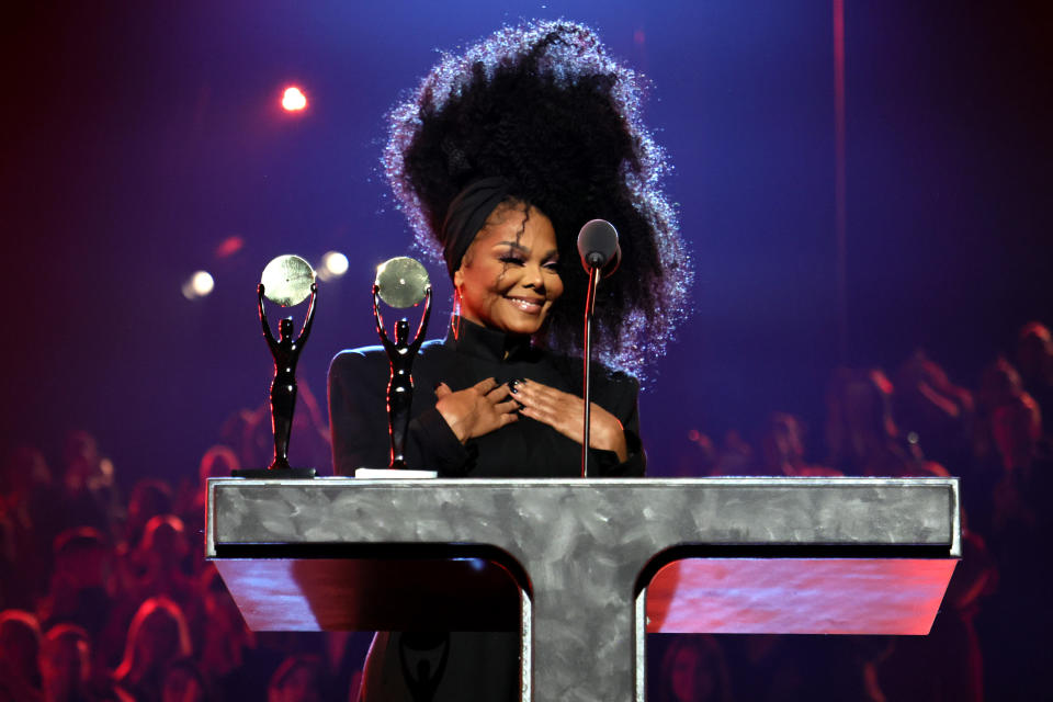 Janet Jackson smiling at the podium in black nostalgic outfit