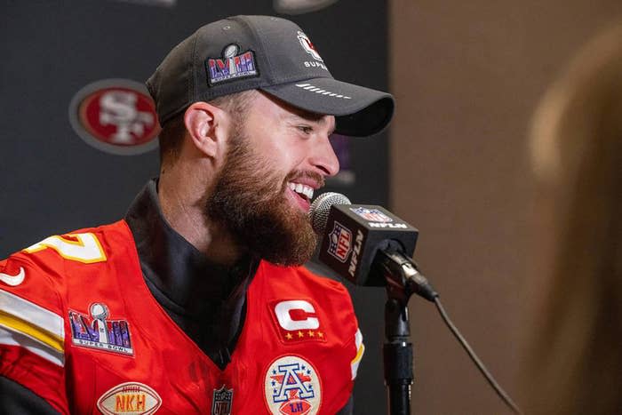 Harrison Butker wearing a sports cap and red football jersey, speaks into a microphone at a press event