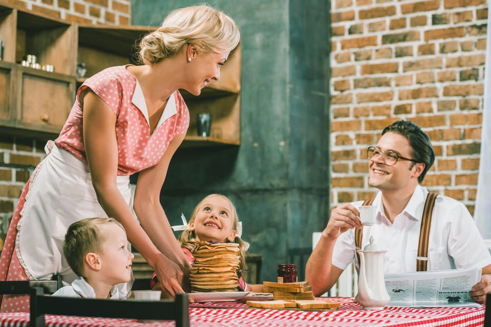 happy 1950s mother holding pancakes for breakfast and looking at husband and kids