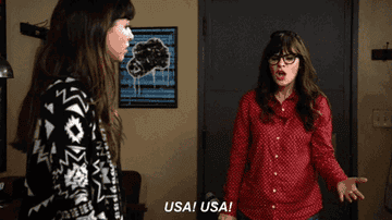 Two people are indoors, one wearing a patterned sweater and the other in a polka dot shirt, saying "USA! USA!"