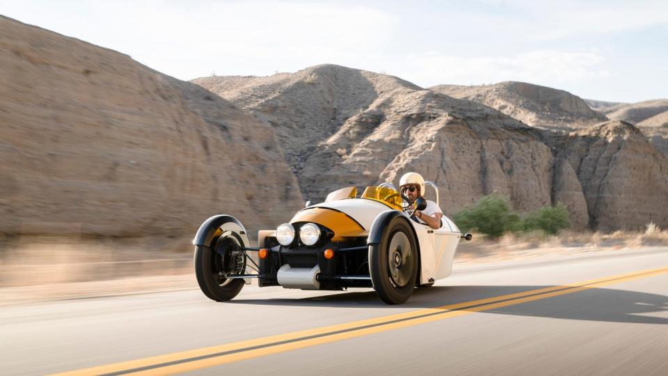 a person driving a yellow 2024 morgan super 3 on a road with mountains in the background