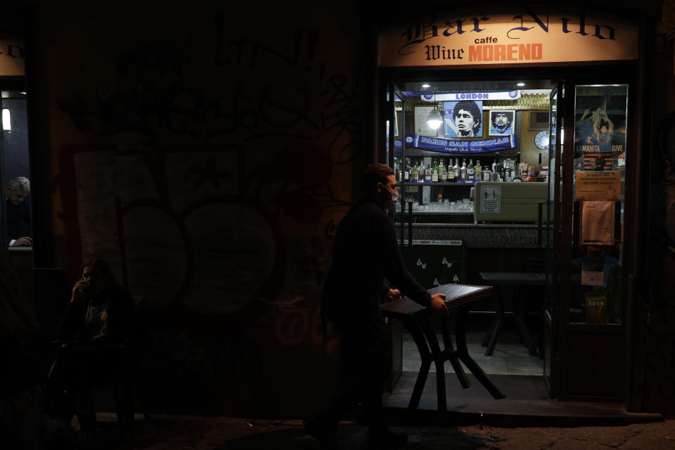 A bartender carries a table inside as he is closing the Nilo Bar in Naples, in the region of Campania, Italy, Friday, Nov. 13, 2020. The regions of Campania and Tuscany were designated red zone on Friday, signaling the dire condition of a hospitals struggling with a surge of new admissions. (AP Photo/Gregorio Borgia)