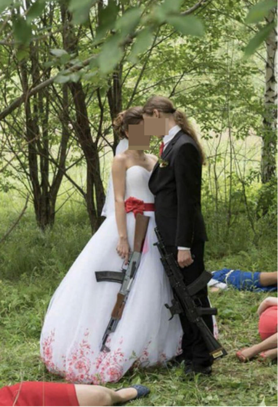 A bride and groom holding assault rifles while people around them lay on the grass