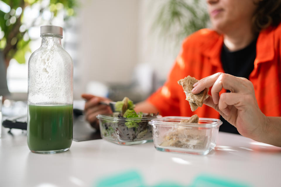 A businesswoman is eating healthy lunch and drinking smoothie while working in her environmentalist green office with lots of house plants and natural light coming from windows.