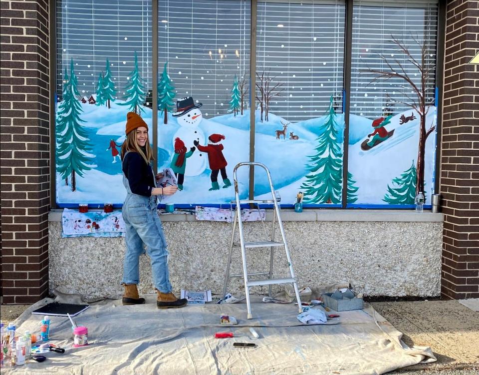 A woman stands next to a wall mural she is painting of a snowy scene outdoors on a window.