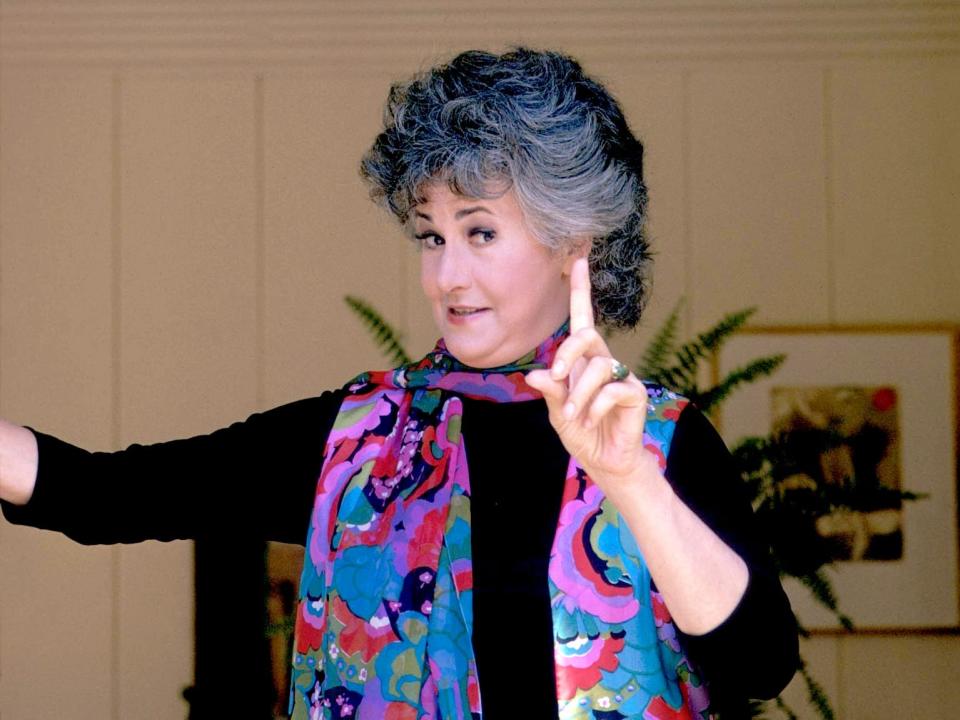 Bea in a colorful blouse and holding up her finger to the camera.