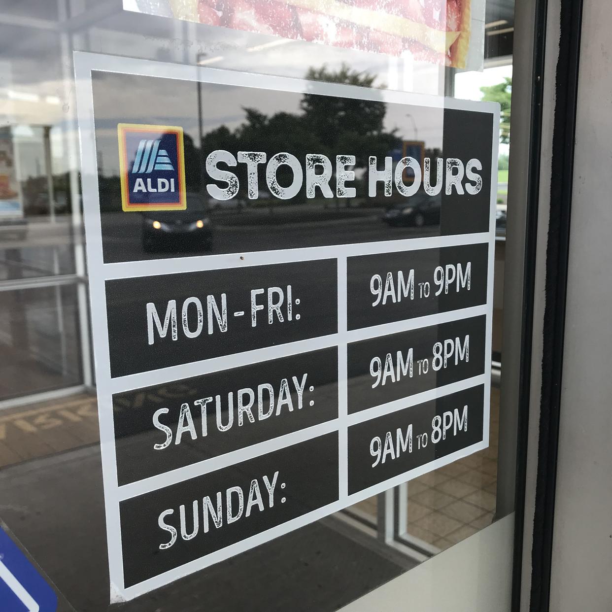 Closeup of a 'Store Hours' sticker on window of an Aldi store, with store hours for 'Mon-Fri', 'Saturday', and 'Sunday', interior front doors entrance in the background through the window