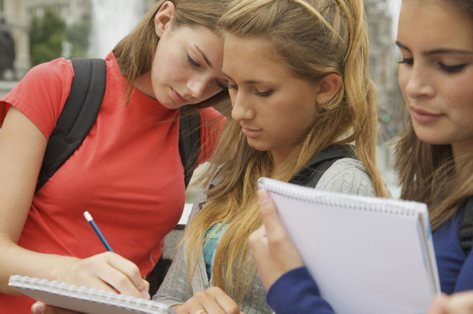 Three young women, wearing casual clothes with backpacks, are focused on writing in notebooks outdoors