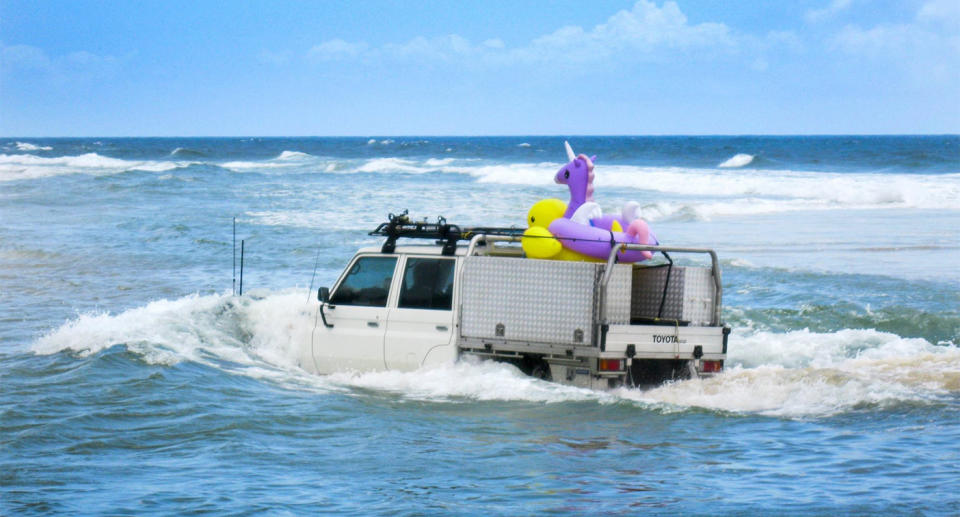 A 4WD drives in water covering the bonnet as it carries inflatable beach toys in the tray.