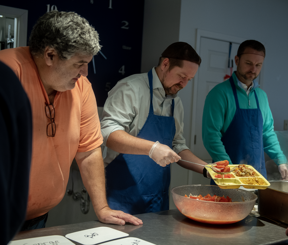 Bill Bowen, Kent Social Services food coordinator, waiting for to-go orders, looks over as Bobby Dalton, national sales manager for Kent Elastomers, scoops a tomato salad onto a tray during a free meal distribution.