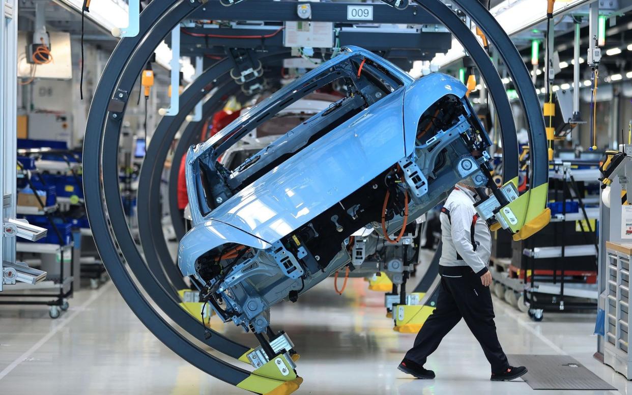 Body shell of an all-electric Porsche Taycan luxury automobile in an overhead conveyor cradle on the production line with man walking behind