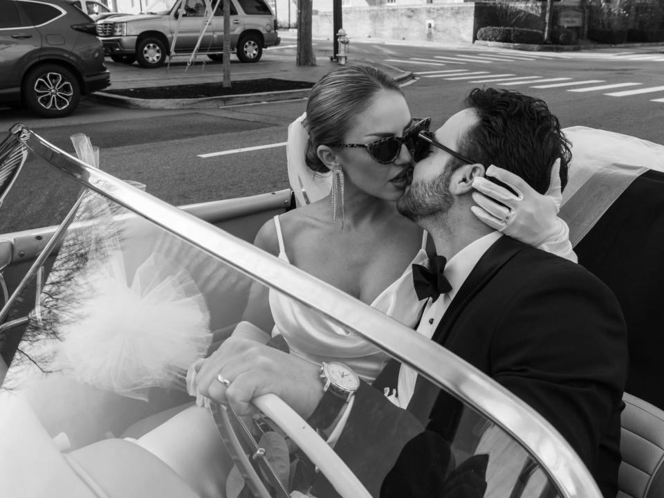 A black and white photo of a woman and man wearing sunglasses and kissing in the car.