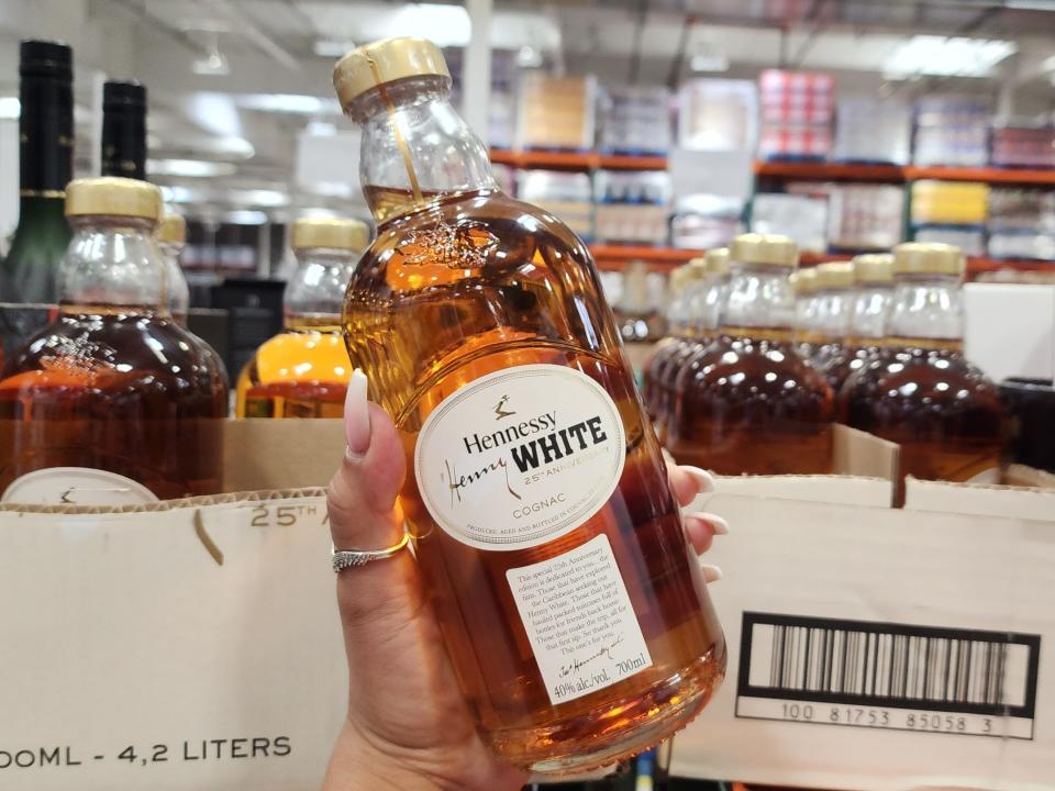 The writer holds a clear bottle of brown Hennessy White in front of other bottles in Costco's liquor department