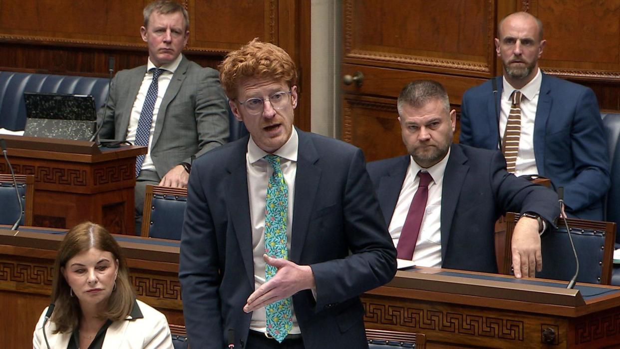 Matthew O'Toole, with red hair, stands in the Assembly chamber wearing round glasses and a navy blue suit with a green tie