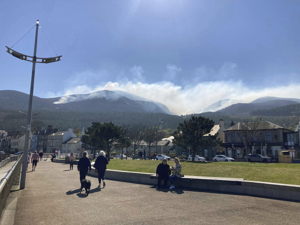 People walk with a view of smoke from the gorse fire spreading across the Mourne Mountains in the background, seen from Newcastle, County Down, in Northern Ireland, Saturday, April 24, 2021. Firefighters in Northern Ireland are spending a second day batting fires through demanding terrain on the Mourne Mountains. Helicopters from both Britain and Ireland are set to join the effort Saturday to put out the fires. They have been raging since Friday morning in the Slieve Donard area — the highest point in Northern Ireland with a peak of 850 meters (2,780 feet). (Rebecca Black/PA via AP)