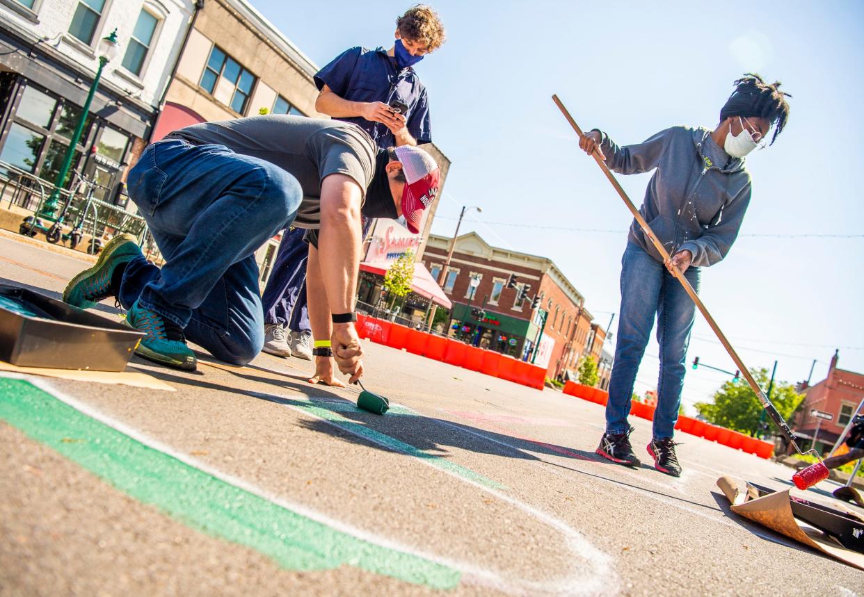 As Sean Starowitz takes a picture with his phone in May 2021, Erik Pearson, left, and Christina Elmore, right, work on the Black Lives Matter mural on Sixth Street in downtown Bloomington.