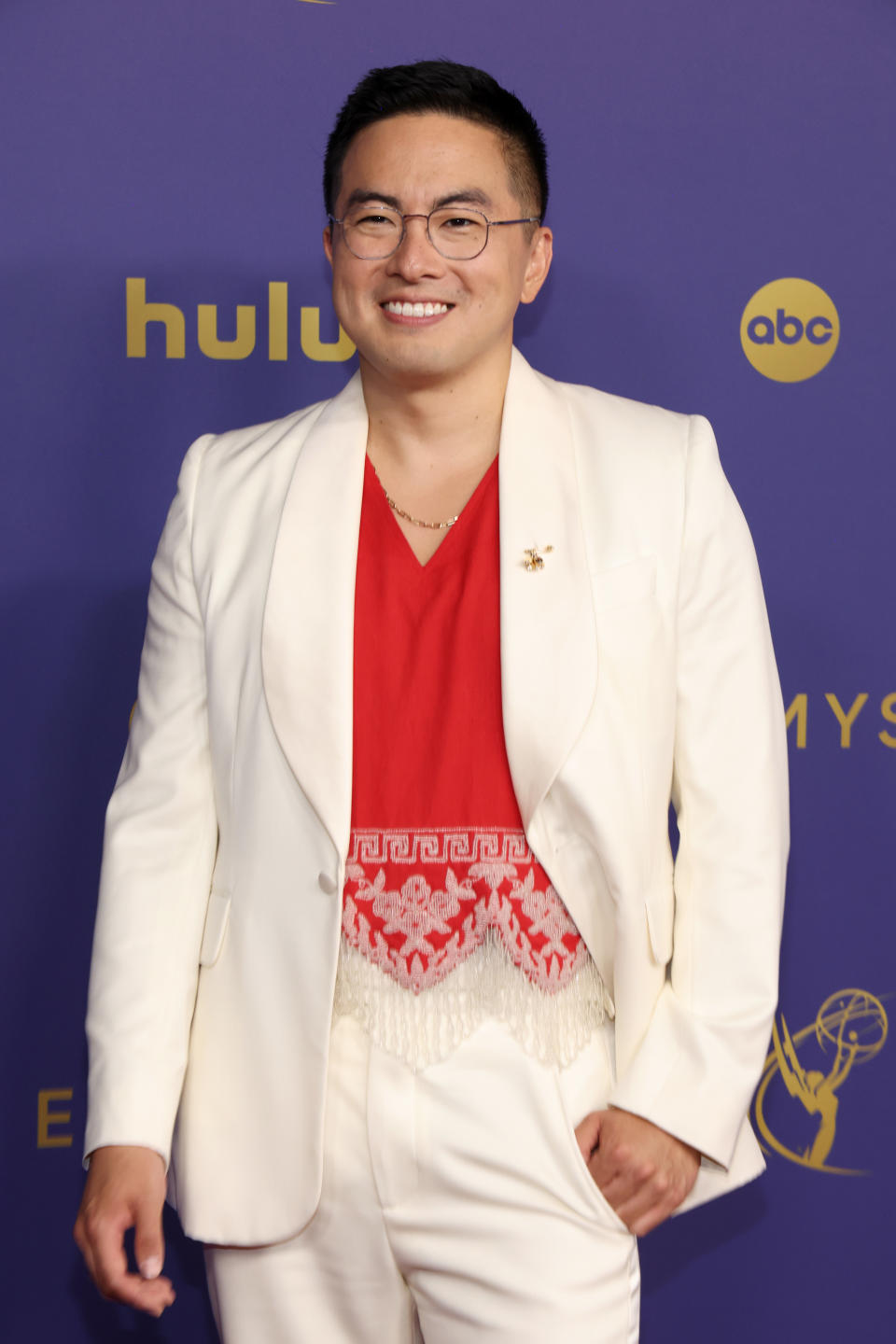 Bowen Yang smiles on the red carpet, wearing a suit with a lace-detailed shirt underneath.