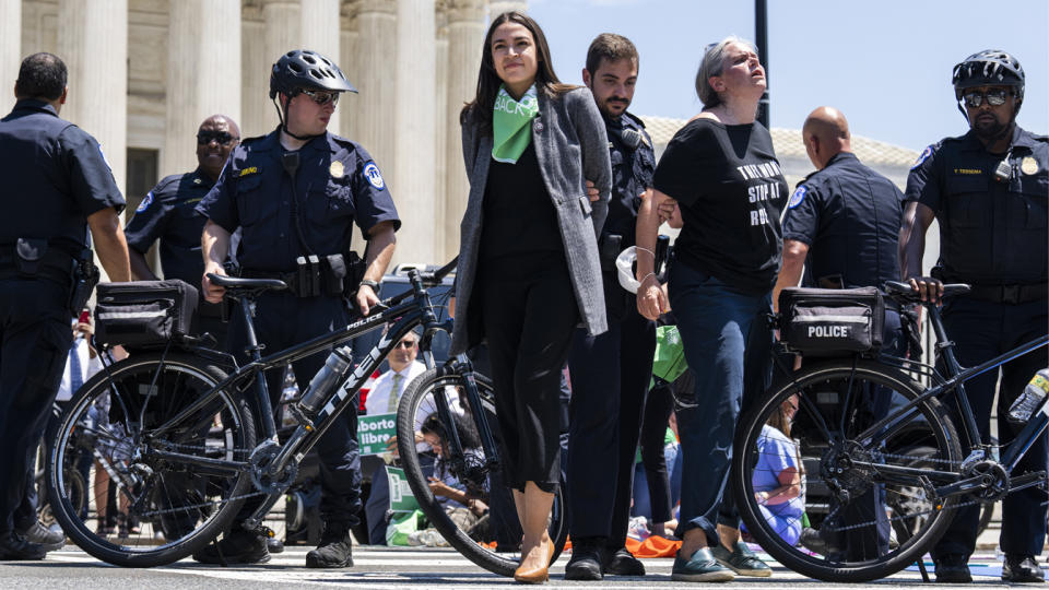 Rep. Alexandria Ocasio-Cortez, D-N.Y., center, looking upbeat, with one hand pulled behind her back by an officer, who holds the arm of another protester who looks thoroughly upset. Five other officers on bicycles stand by.