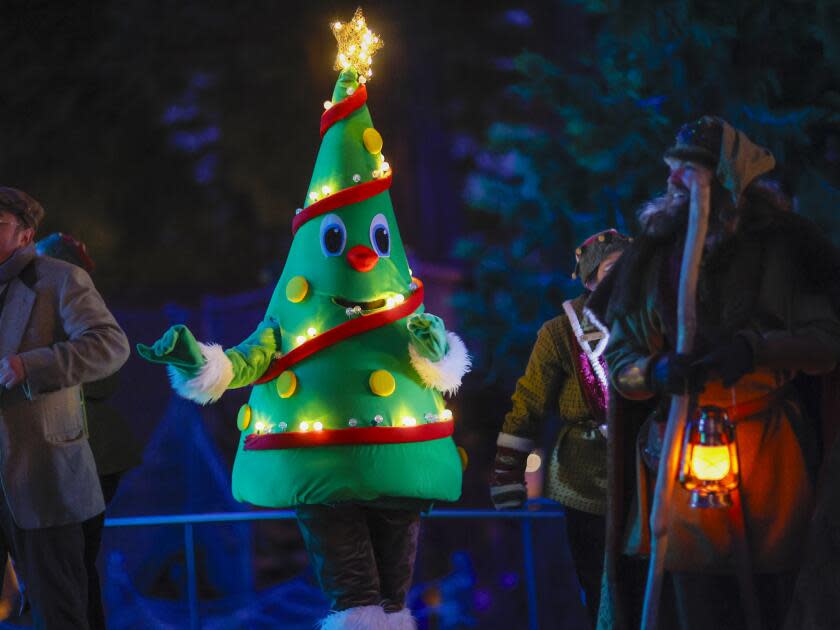 A person in a Christmas tree costume walking around a Christmas village at night.