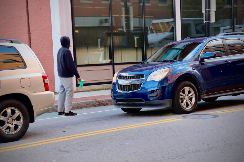 A man stands on the side of Centre Street in Brockton as cars drive by.